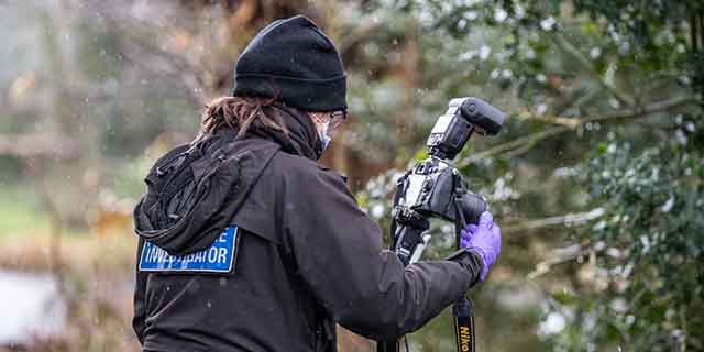 Crime scene investigator wearing gloves and outdoor gear, adjusting a camera on a tripod in a wooded area.