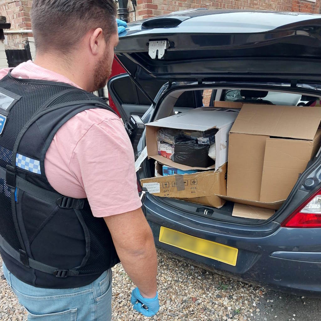 Plain clothed police officer wearing body armour looks into a loaded car boot