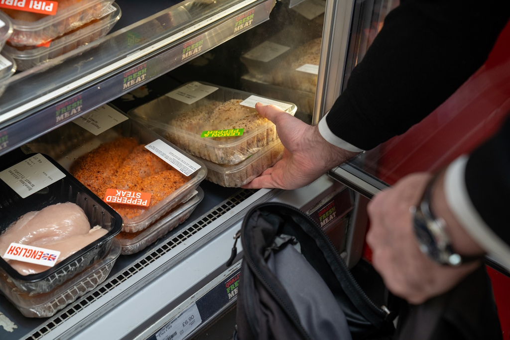 A hand lifts a pack of pepper and lemon chicken breasts  out of a chiller cabinet, ready to place it in an open rucksack