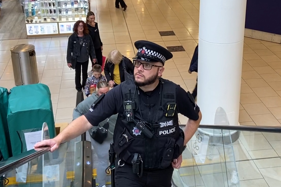 A uniformed Essex Police officers rides up an escalator in a busy shopping centre