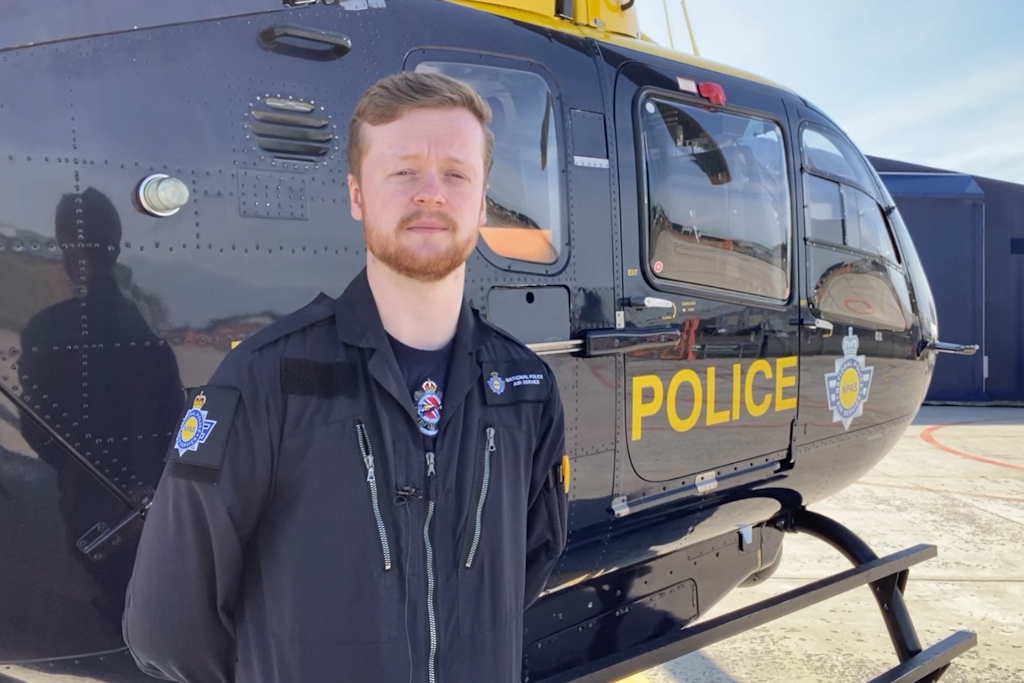A police officer in police helicopter flight crew overalls stands in front of a yellow and blue National Police Air Service liveried helicopter