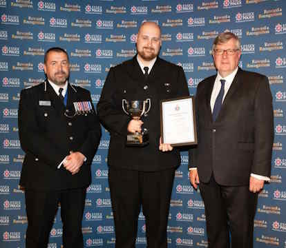 Three men stand in front of a logo backdrop; the man in the center holds a trophy and certificate, flanked by a uniformed officer and a man in a suit