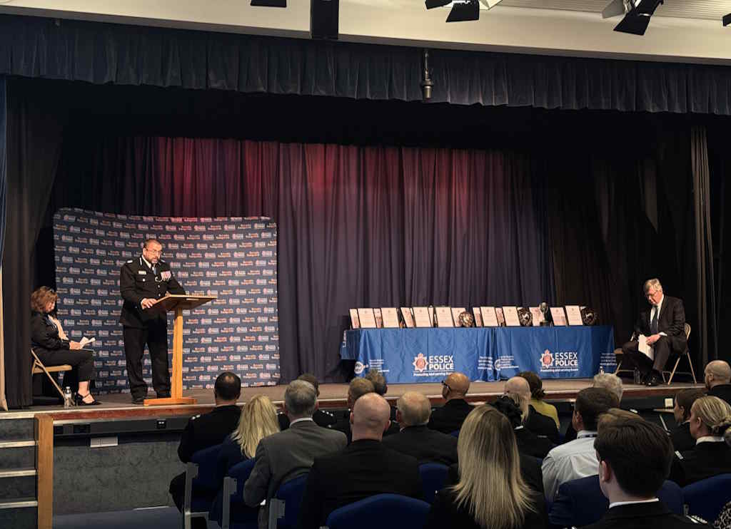 A police officer speaks at a podium on a stage with an 'Essex Police' backdrop, flanked by two seated individuals. A table with awards is on the stage, and the audience is seated facing them.