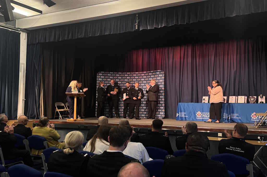 A group of people in uniform stand on a stage receiving awards, with an audience seated in front. The backdrop displays 'Essex Police' logos, and a table with more awards is visible to the right.