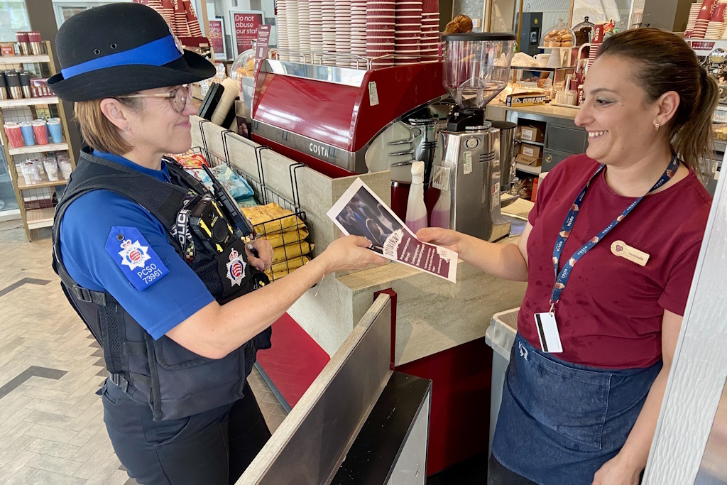 A uniformed Essex Police police community support officer hands an explanatory leaflet about spotting the signs of child exploitation to a member of staff in a coffee shop at a rail station