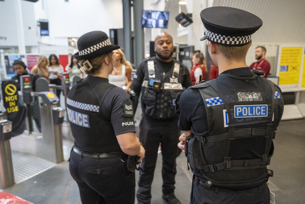 Two uniformed Essex Police officers speak to a member of Greater Anglia railways staff at a busy rail station