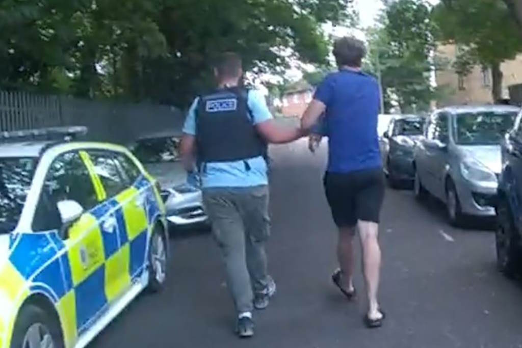 A plain-clothes police officer leads a suspect away to a parked police car