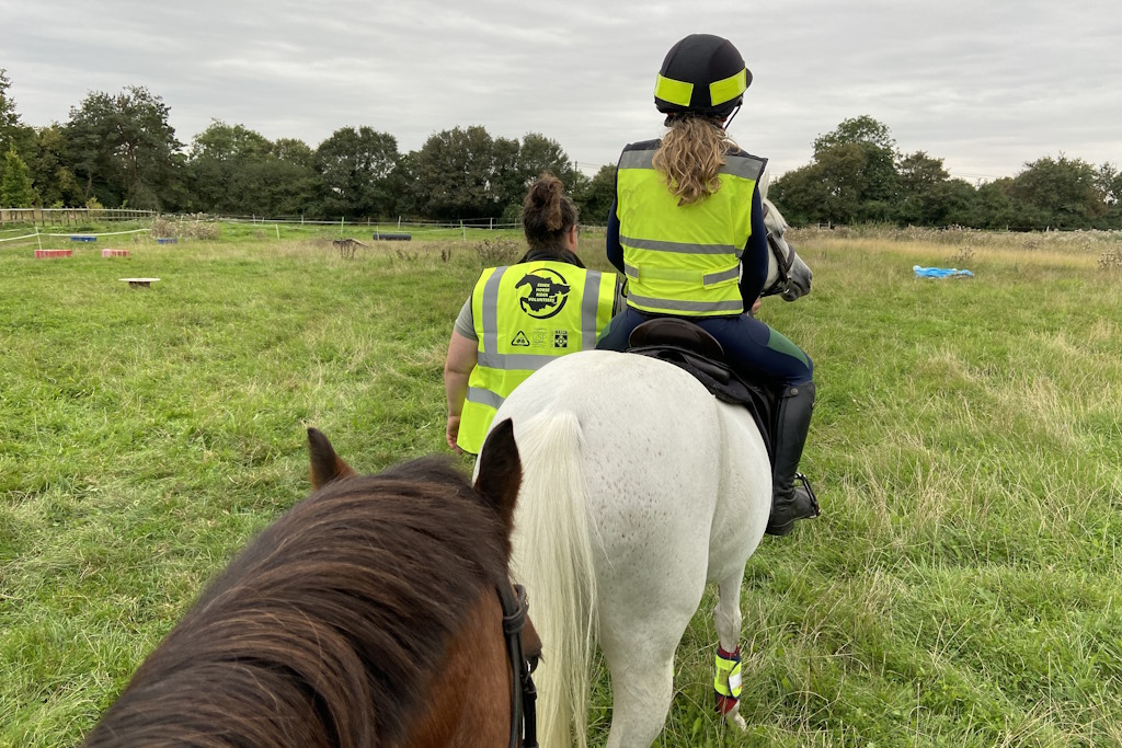 The young rider of a white horse is accompanied on a ride across a field by a woman wearing a yellow fluorescent vest with the Essex Horse Rider Volunteer Scheme logo and words printed on the back