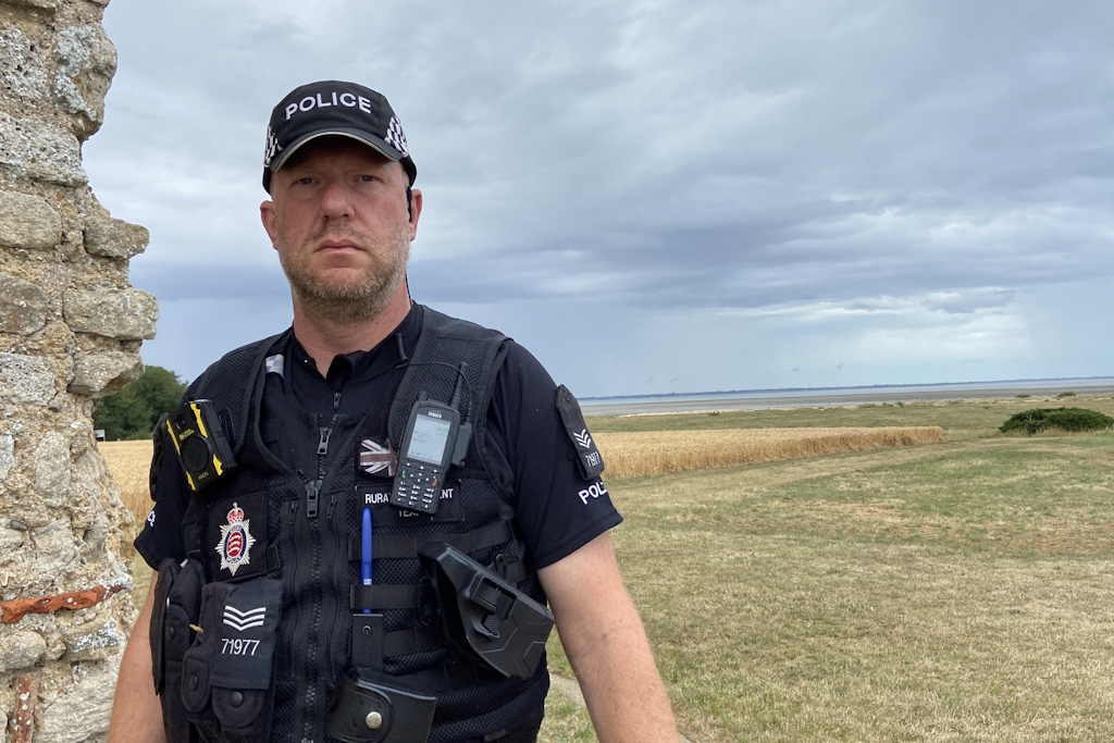 An Essex Police rural engagement officer walks past the stone and brick wall of an ancient church, with fields and the sea in the distance, beneath a blue sky