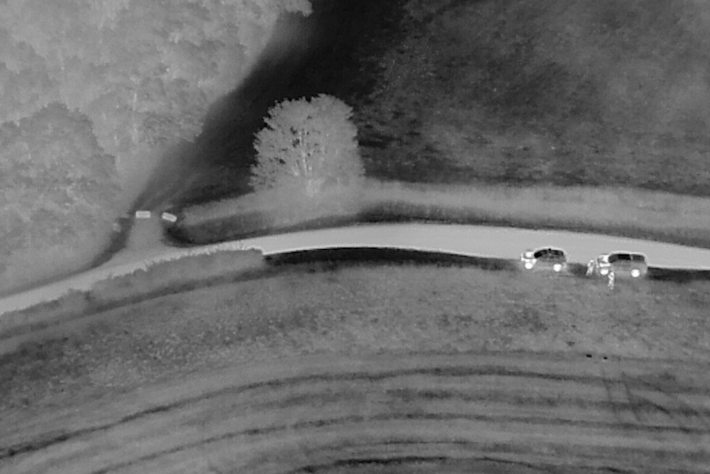 An aerial infra-red image from a drone showing two vehicles and two people next to a lane by the side of a field
