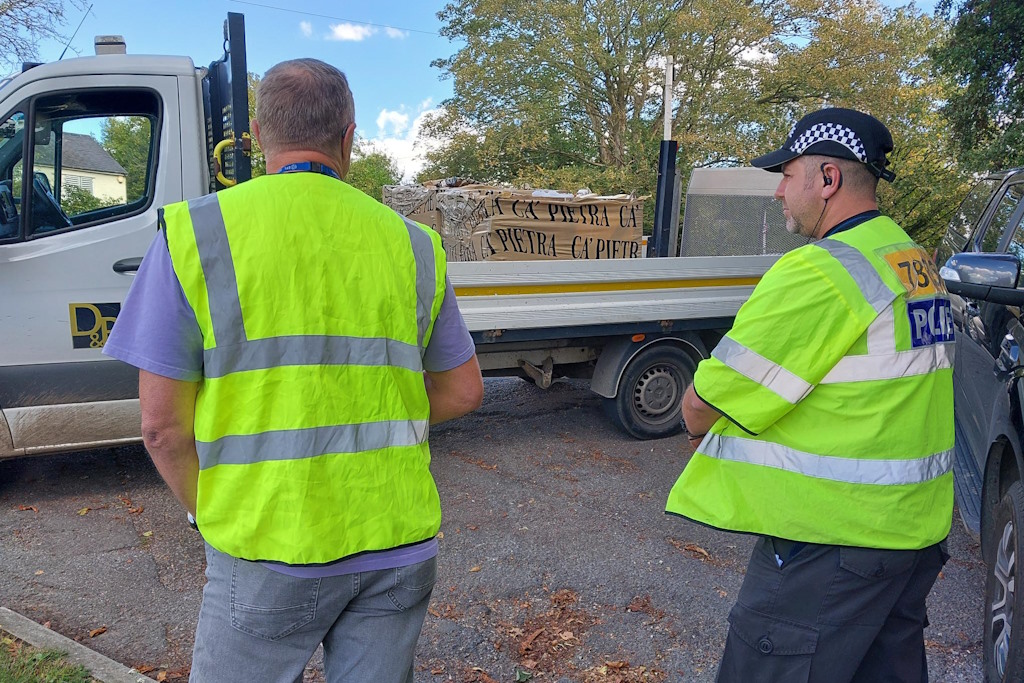 A uniformed Essex Police rural engagement officer and a member of council staff, both wearing yellow fluorescent jackets, speak to the driver of a flatbed lorry loaded with building materials