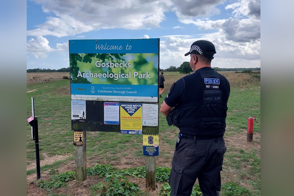 A uniformed Essex Police rural engagement officer pauses by a large sign proclaiming Welcome to Gosbecks Archaeological Park