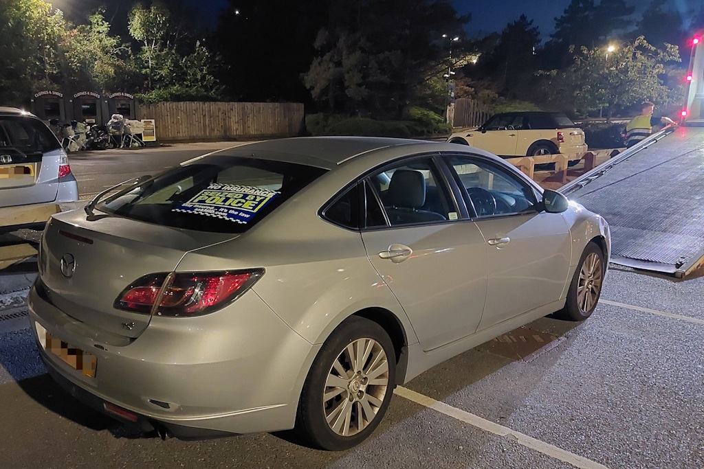 A grey car being winched on to the back of a trailer after having been seized under section 165 of the Road Traffic Act. A sign in the back window reads No insurance. Seized by police. Stay insured. It's the law.