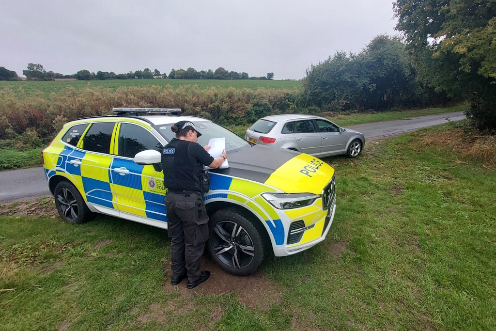 A uniformed Essex Police rural engagement officer leaning on the bonnet of a liveried police vehicle completing paperwork to seize a silver car parked on a rural lane behind them