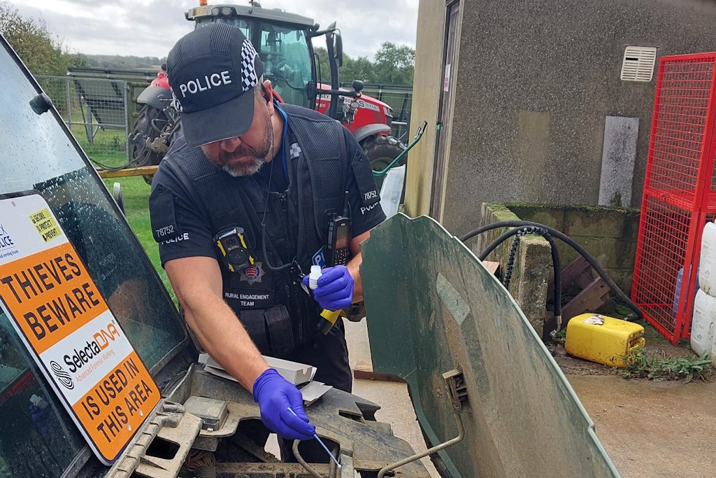 A uniformed Essex Police officer, wearing purple latex gloves, uses a wand to mark a farm vehicle with a special DNA solution. A sign on the vehicle's windscreen states: Thieves beware. Selecta DNA is used in this area. There is a tractor in the background