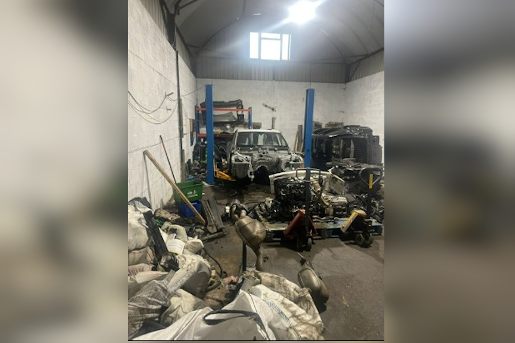 Cluttered workshop with dismantled car on a lift, surrounded by scattered engines, exhausts, and parts.