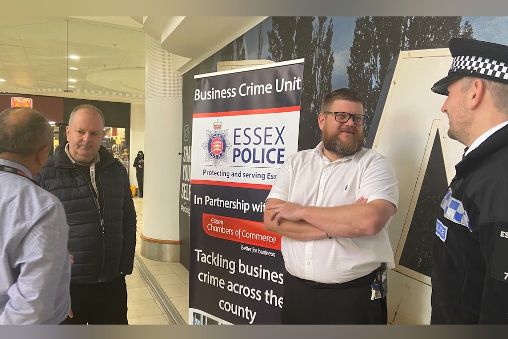 A group of people, including a uniformed Essex Police officer and a business crime officer in plain clothes chat in front of a banner in a shopping centre. The banner is advertising the Essex Police Business Crime Team 