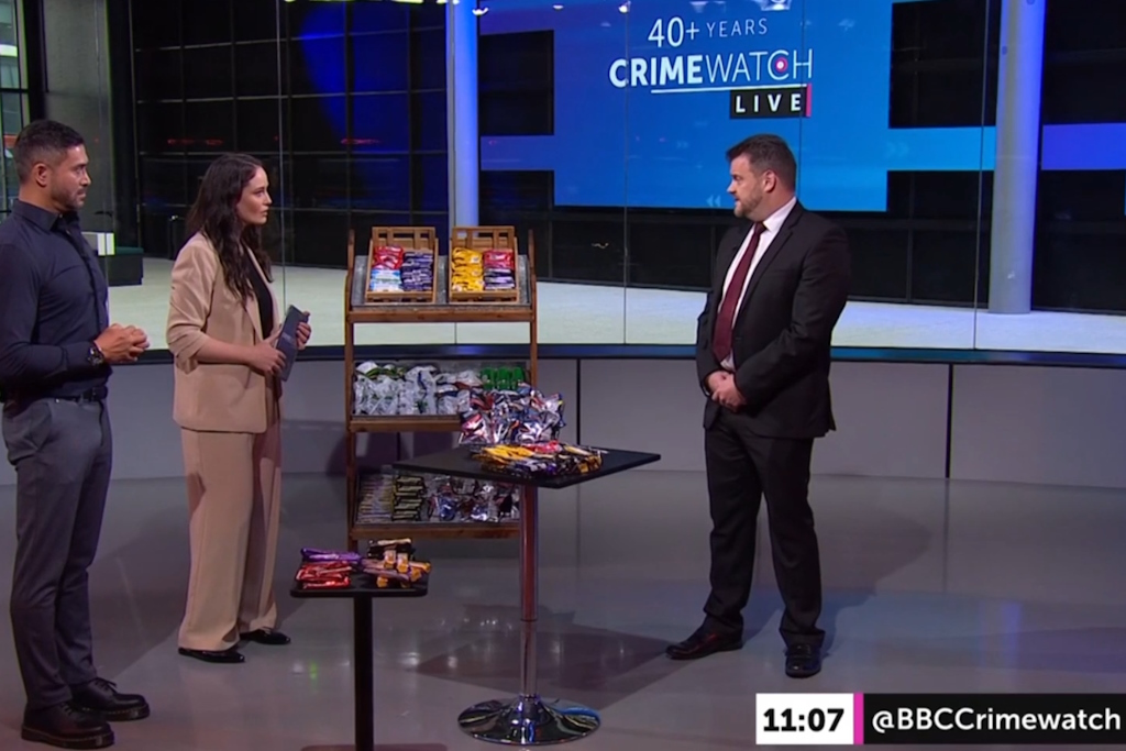 An Essex Police Business Crime Team officer in plain clothes speaks with a BBC presenter on the Crimewatch Live set, surrounded with shop goods displayed on shelves and two tables. A screen in the background announces 40+ years Crimewatch Live