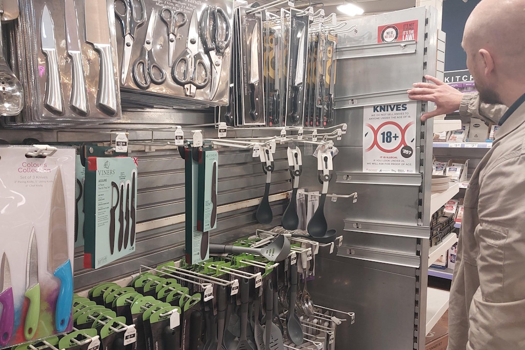 An Essex Police business crime officer in plain clothes checks a shop display of knives and sharp implements. He is reading a Challenge 25 poster warning purchasers that they will be challenged for proof of age if they appear to be under 25