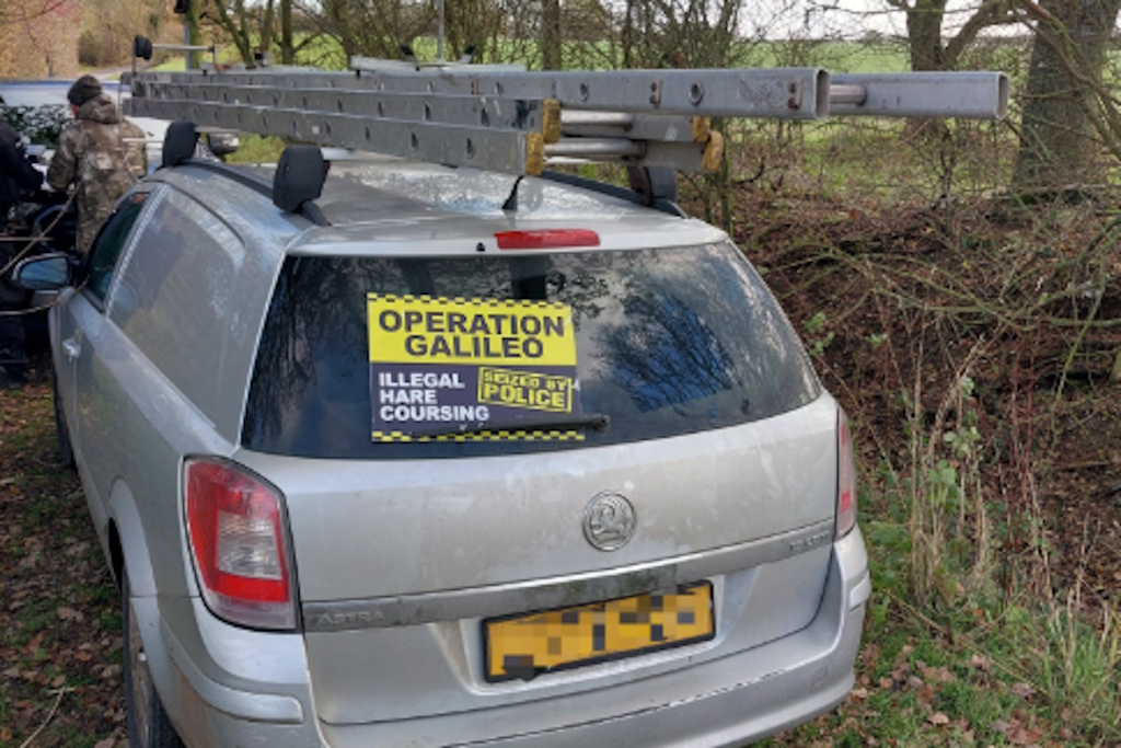 A silver car with a ladder on its roofrack has a blue and yellow sign on its back window, which states: Operation Galileo. Illegal hare coursing. Seized by police. in the background a police officer is speaking to a man in a khaki camouflage jacket, who is signing a document while leaning on the back of another vehicle, parked in a rural lane next to a row of trees