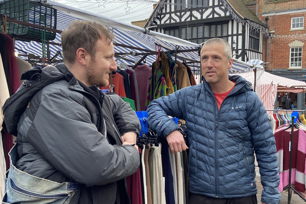 Two men lean on a rail of skirts in front of a covered market stall with rails of coats and knitwear