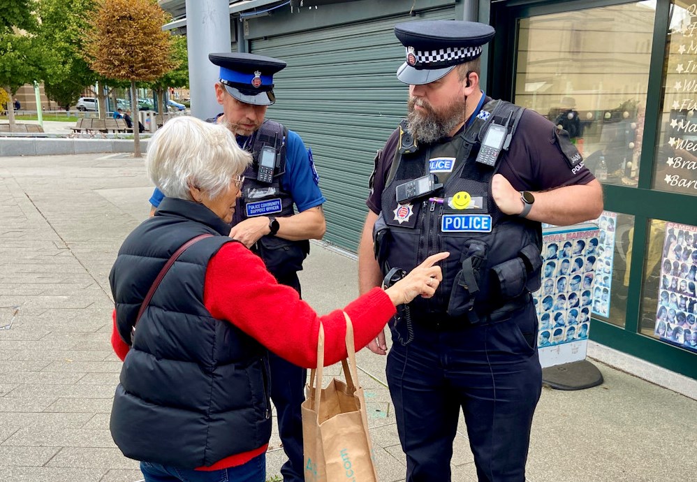 Two police officers in uniform speak to a grey-haired woman while on patrol