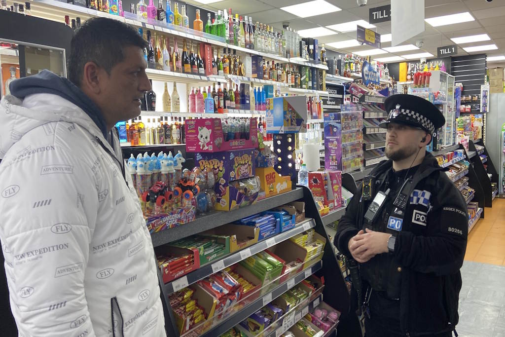 A uniformed police officer stands in a shop speaking with a man in a white coat