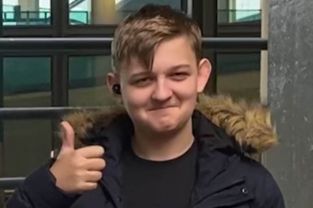 A smiling teenage boy with light hair and a hooded dark coat with his thumb up
