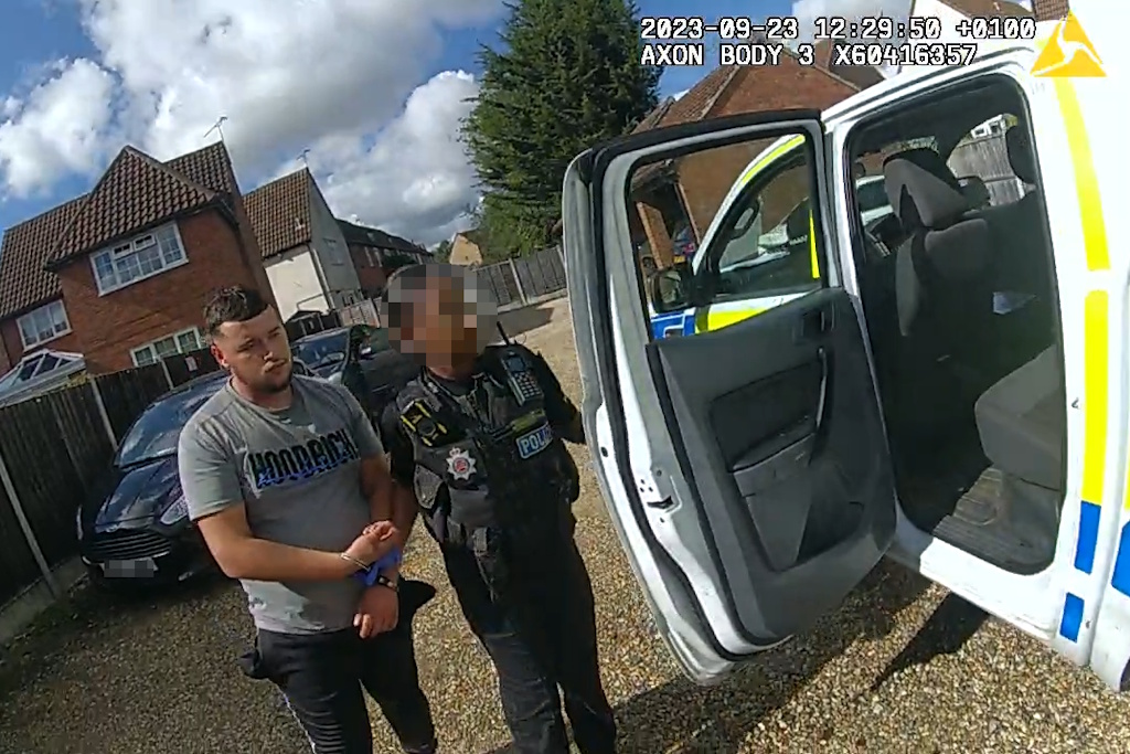 Essex Police body-worn video still showing a man in handcuffs being led to the open door of an Essex Police liveried vehicle by a uniformed Essex Police neighbourhood policing officer