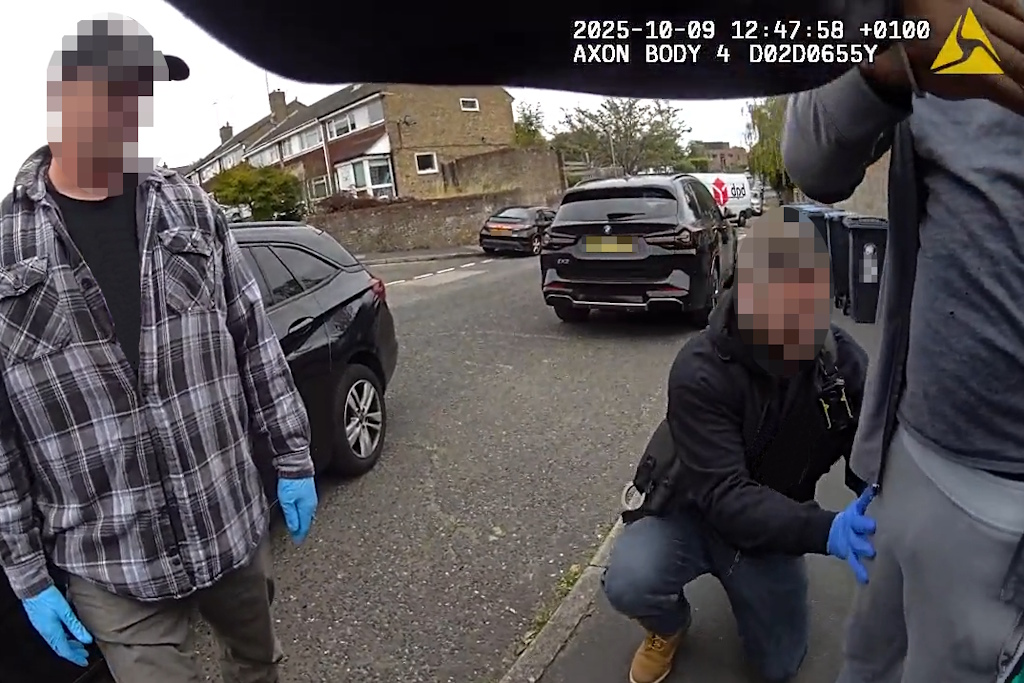 Essex Police body-worn video footage of a police officer in plain clothes searching a man wearing a grey tracksuit, watched by another officer in plain clothes. Both are wearing blue safety gloves