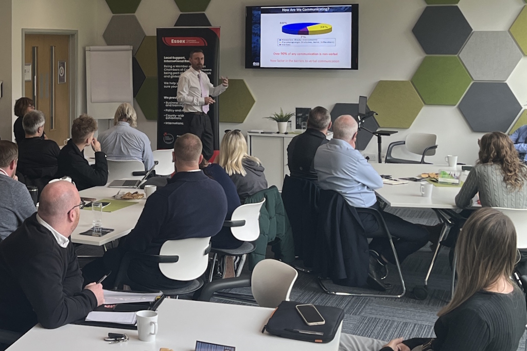 A group of men and women sitting at tables facing a man who is talking in front of a screen on the wall which contains a coloured pie-chart below the question How are we communicating? The slide states that more than 90% of any communication is non-verbal