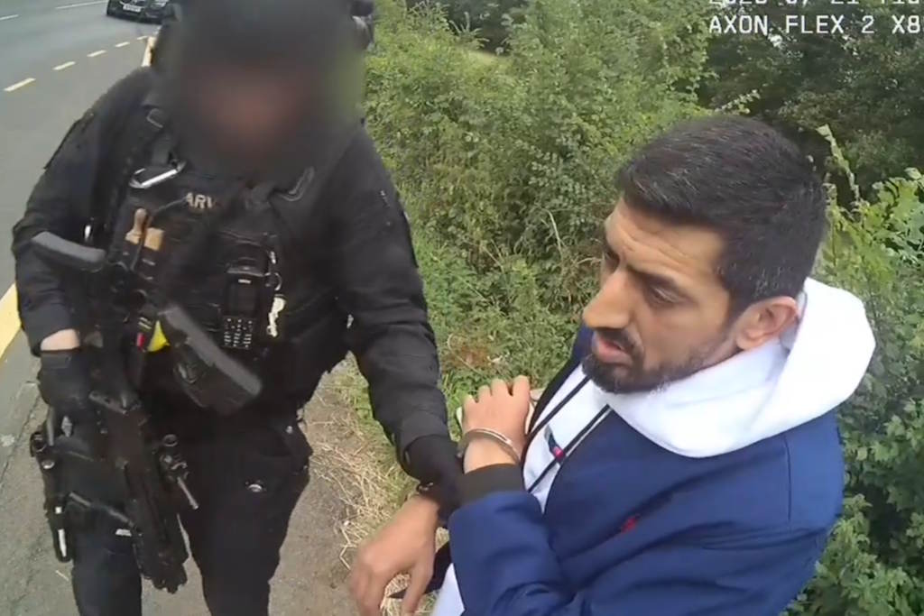 A uniformed armed officer holding a set of handcuffs, attached to a bearded man wearing a white hoodie and blue jacket