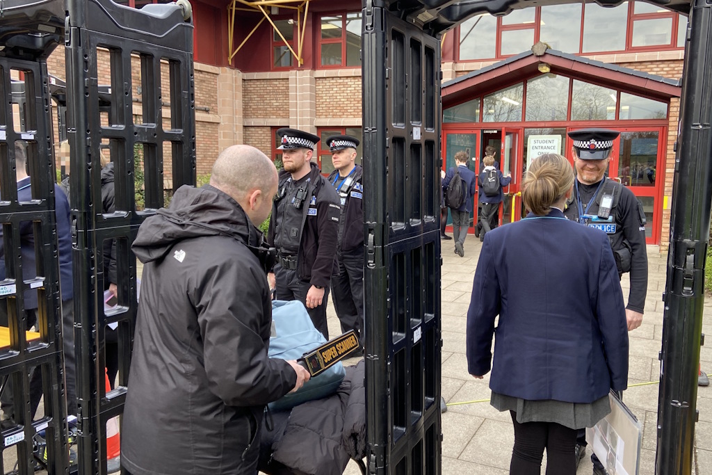 A school pupil wearing a blue blazer walks through a knife safety arch, watched by a uniformed Essex Police neighbourhood policing officer. Beside the arch, a teacher scans a school bag with a security wand. A brick school building, with pupils entering, stands in the background