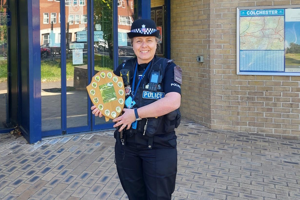 A uniformed Colchester neighbourhood policing officer stands in front of a police station holding a shield