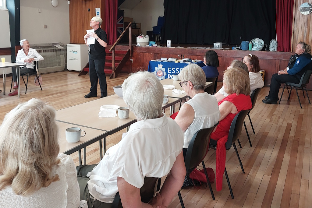 A uniformed Essex Police staff member speaks to an audience seated at tables in a hall, with an Essex Police banner visible