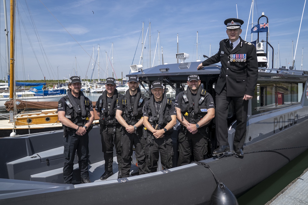 Five uniformed Essex Police Marine Unit officers stand aboard a police launch in a marina next to the Essex Chief Constable in ceremonial uniform