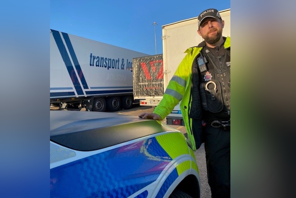 A uniformed Essex Police rural engagement officer rests his hand on the bonnet of a liveried Essex Police vehicle. He is standing in front of a number of lorries queueing up to load on to a North Sea ferry