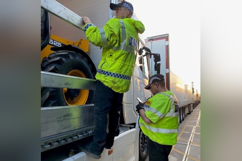 A uniformed Essex Police rural engagement officer and an officer from the National Construction and Agricultural Theft Team, both wearing fluorescent jackets, search a lorry with a digger on board. A queue of lorries stretches into the distance