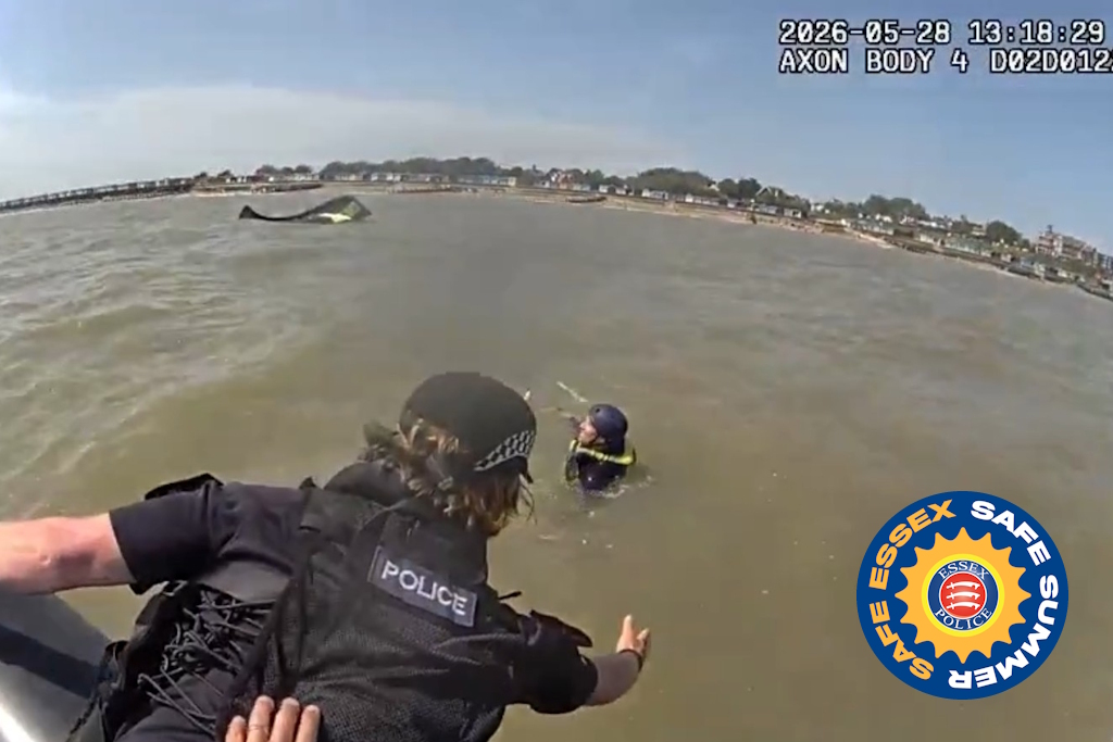 Essex Police body-worn video still of a uniformed marine officer leaning into the water. His outstretched arm is reaching towards a person in the water. in the distance is a board, with its kite lying in the water. There is a beach and houses in the background. The image includes an Essex Police logo, stating Safe Essex, Safe Summer