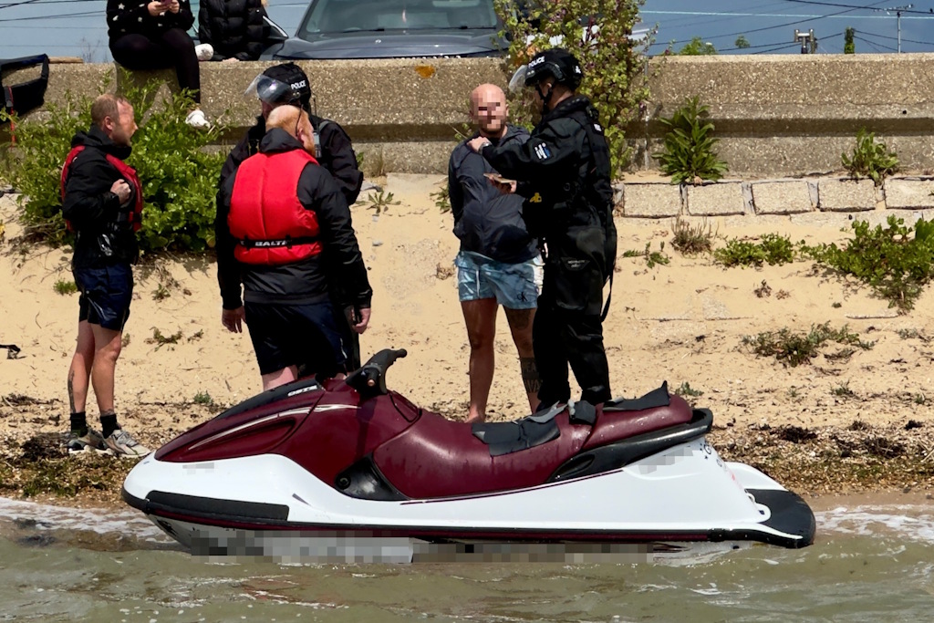 Two uniformed Essex Police marine officers speaking with three men on a beach next to a personal watercraft in the water