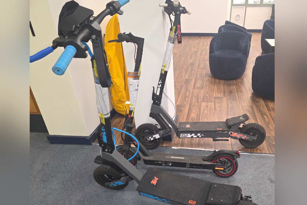 Three grey and black electric scooters, parked indoors on a wooden floor. Blue grips on one of the scooters