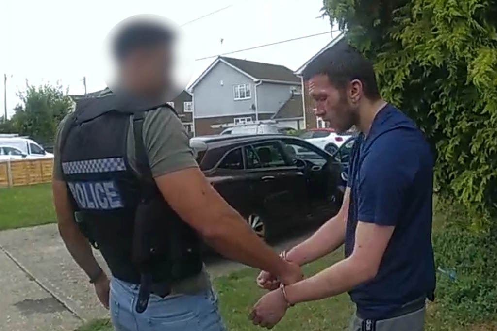 A uniformed officer holds a set of handcuffs, worn by a man in a blue T-shirt
