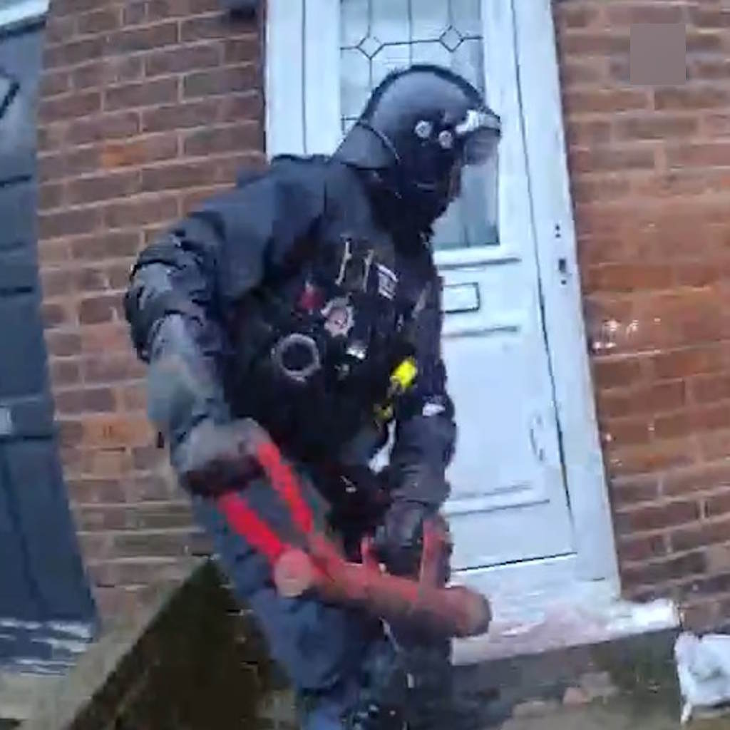 An officer in full kit swings a red ram towards a white front door