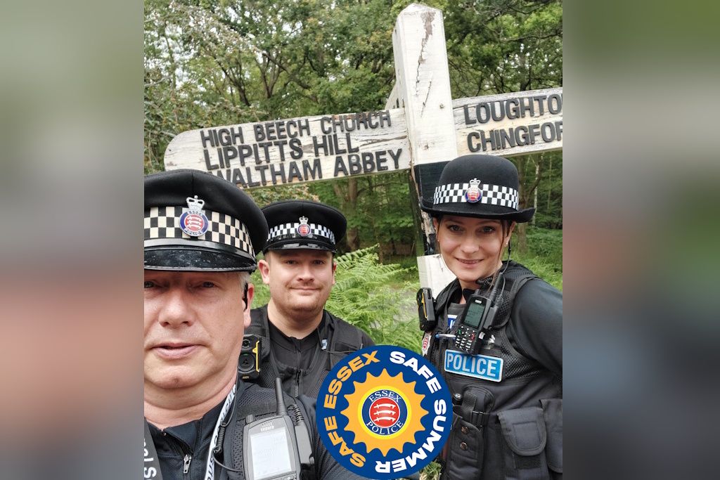 Three uniformed Essex Police neighbourhood policing officers stand next to a wooden fingerpost sign in a forest. The sign points one way to High Beech Church, Lippitts Hill and Waltham Abbey. It points to Loughton and Chingford in the other direction