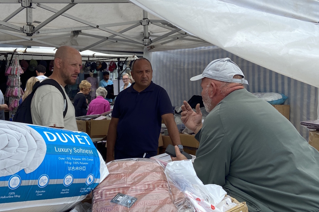 A market stall holder speaks to two plain clothes Essex Police business crime officers across a stall stacked with duvets and bedding
