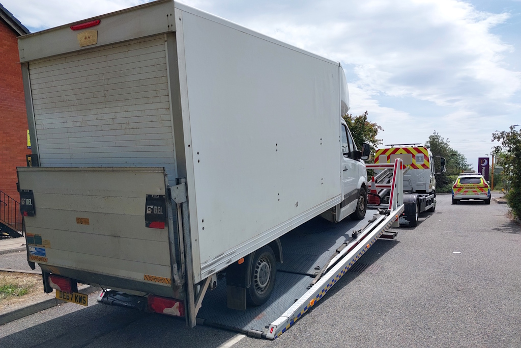 A large van is loaded onto the back of a recovery lorry. A liveried Essex Police vehicle is in the background