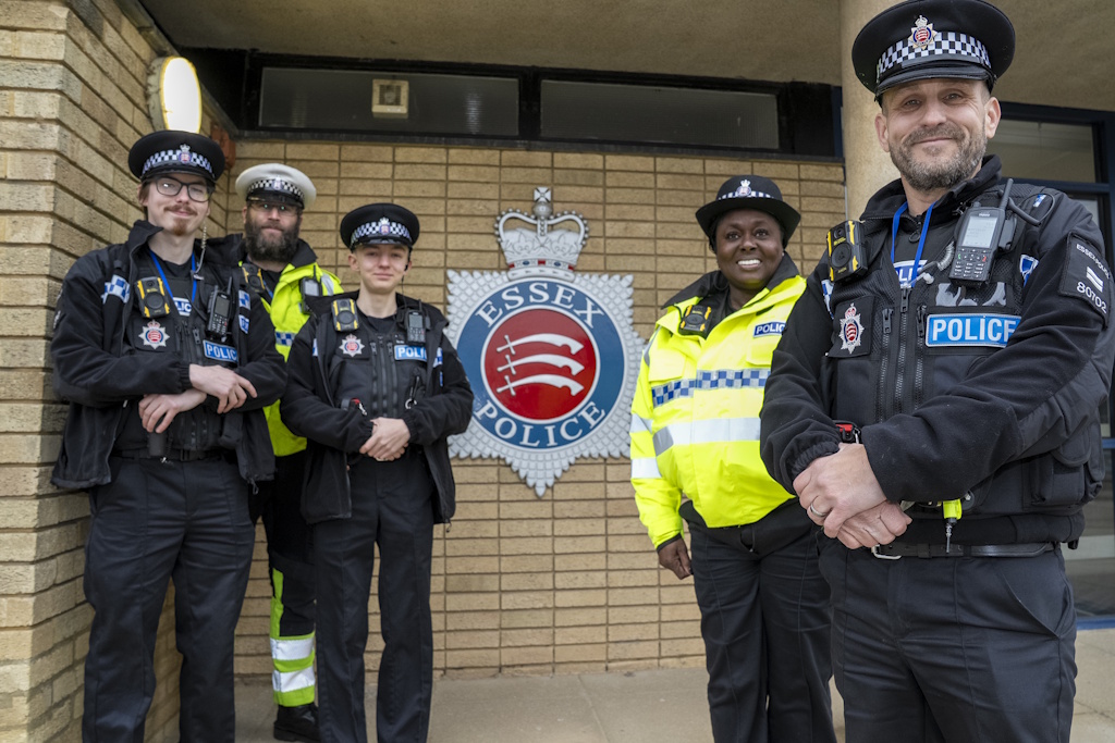A group of five uniformed Essex Police Special constabulary officers standing in front of a large Essex Police crest on a wall. One is wearing a roads policing uniform of white peaked cap, yellow fluorescent jacket and trousers with yellow fluorescent panels. Another officer is wearing a yellow fluorescent jacket