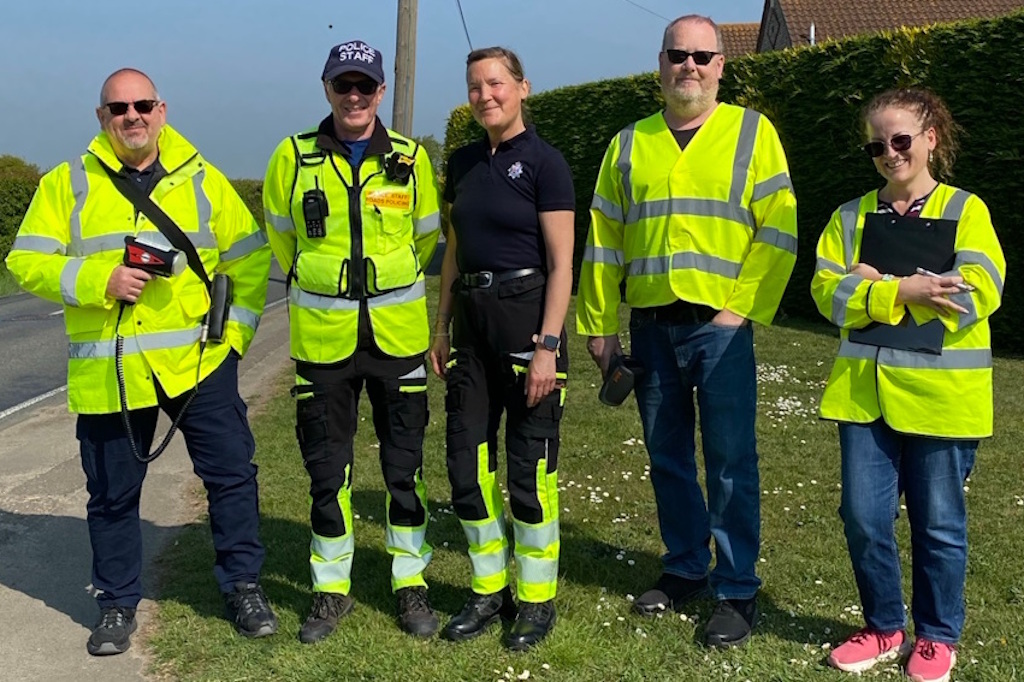 Three community speedwatch volunteers wearing yellow fluorescent jackets, two are carrying speed detection devices, another is carrying a clipboard. They are accompanied by a member of Essex Police police staff and an Essex Police Special officer wearing trousers with fluorescent panels, designed for roads policing officers. Both are in uniform and the police staff member is also wearing a yellow fluorescent jacket