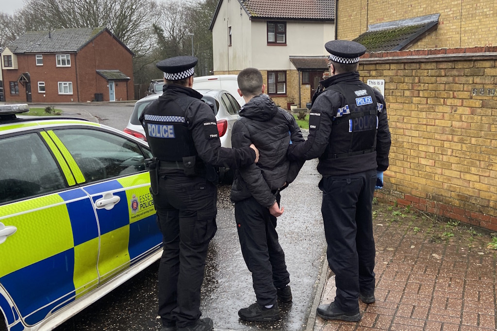 Two uniformed Essex Police neighbourhood policing officers stand with a man they have arrested next to a liveried police car on a housing estate
