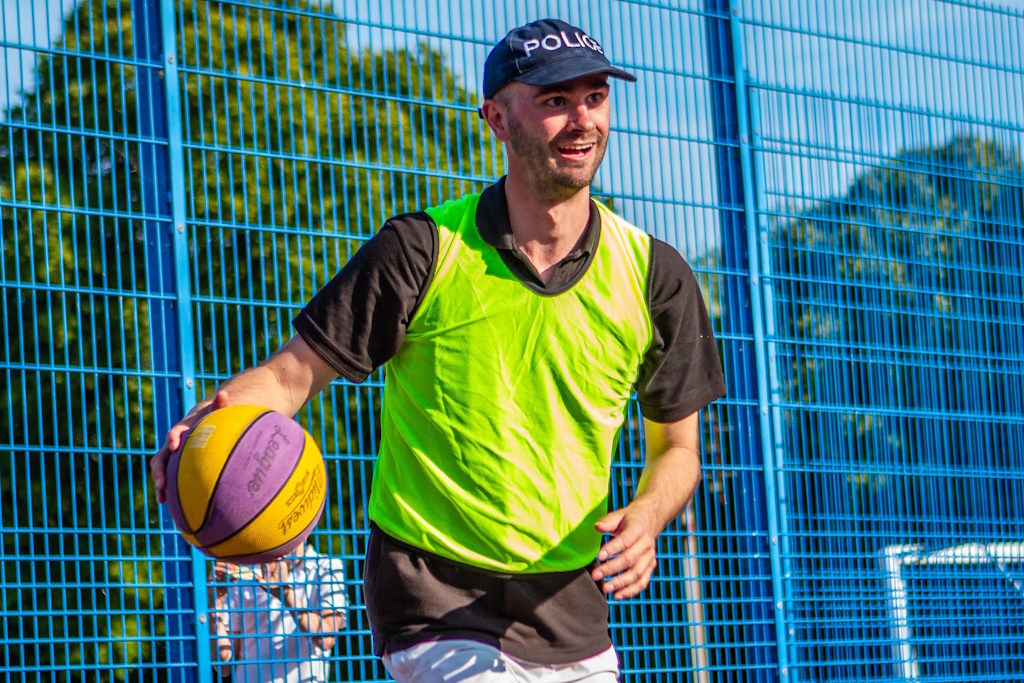 A Colchester neighbourhood policing officer, wearing a police cap and a fluorescent yellow bib, dribbles a yellow and purple basketball in front of a blue mesh fence